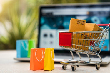 Carrito de supermercado con bolsas y cajas de compras con un computador portatil detrás.